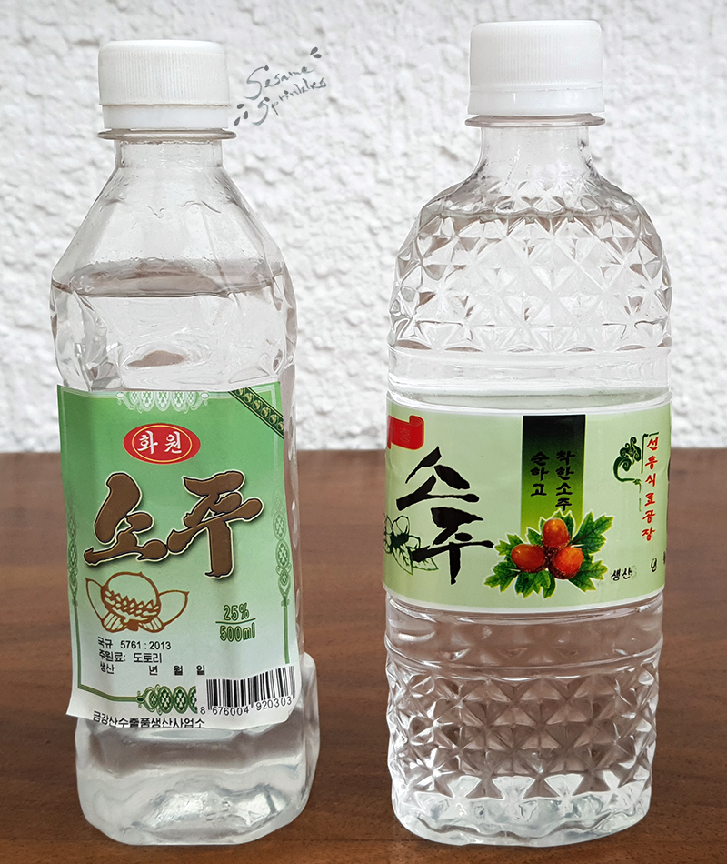 Clear liquor in clear bottles with green labels. The labels on the two bottles are decorated with images of acorns and say "Soju" in font that it typical for North Korea.
