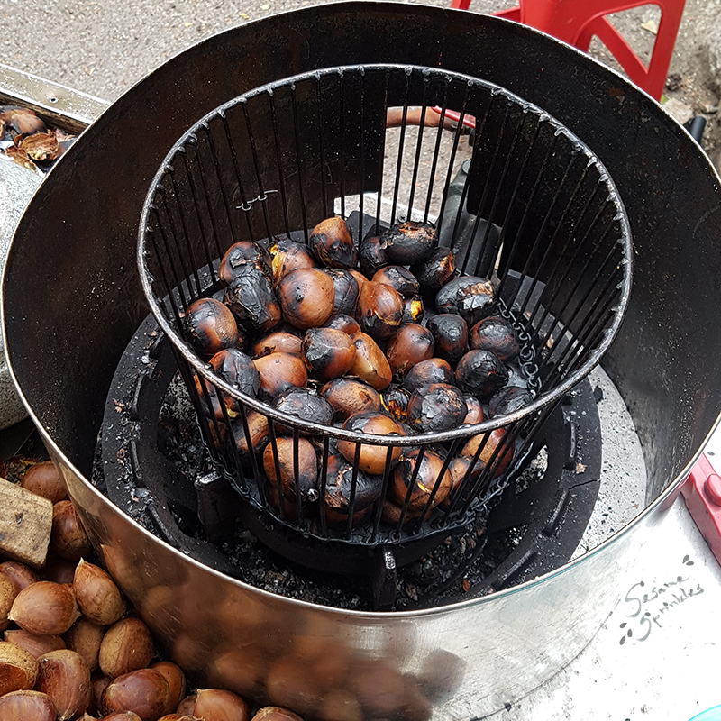 Whole chestnuts inside a metal basket are being roasted over a fire. The skin of most of the chestnuts is partially burnt and cracked, golden yellow flesh is visible through some cracks.
