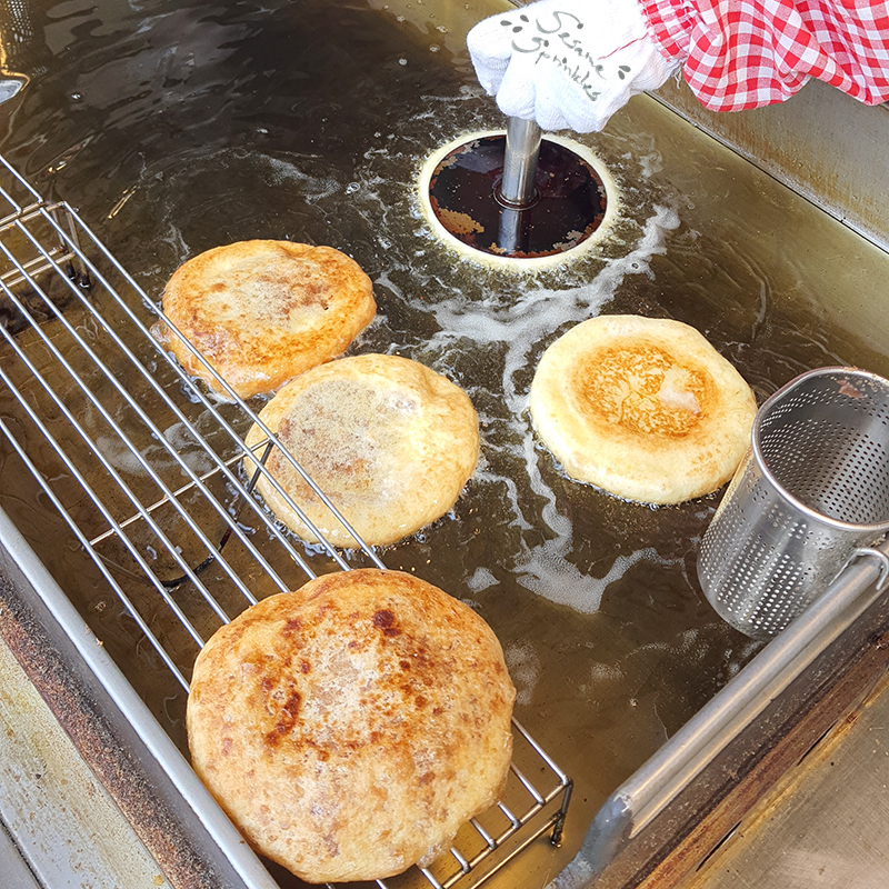 Hotteok being made fresh. One pancake is done and rests on a metal grid. Four pancakes are frying in oil, one of them being pressed down with a flat tool.